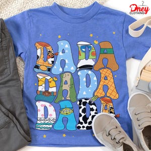 May include: A light blue t-shirt with the word "DADA" in large, colorful letters. Each letter is designed with a different pattern and character theme. The shirt is paired with gray pants and beige shoes.