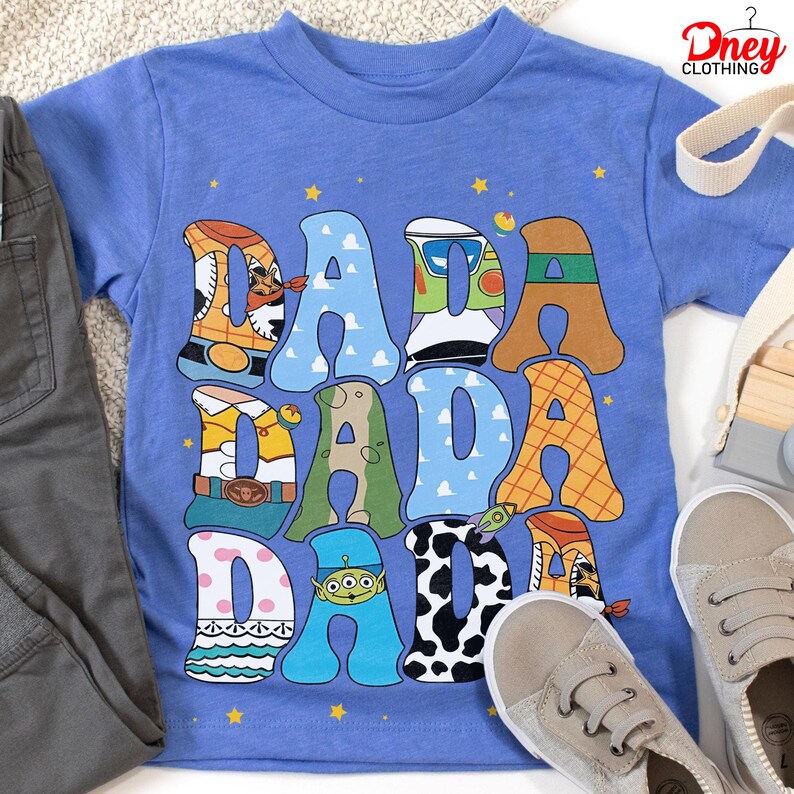 May include: A light blue t-shirt with the word "DADA" in large, colorful letters. Each letter is designed with a different pattern and character theme. The shirt is paired with gray pants and beige shoes.