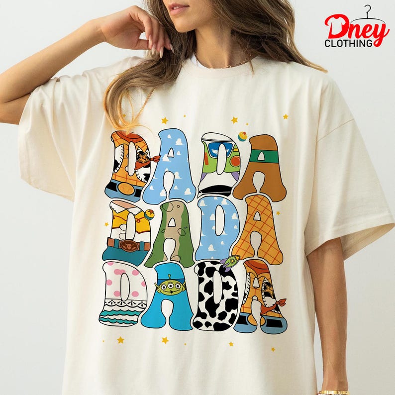 May include: Cream-colored t-shirt with the word "DADA" in colorful, cartoon-style letters. Each letter is decorated with different patterns and characters, including a cowboy, a giraffe, and an alien. The shirt has a relaxed fit and short sleeves.