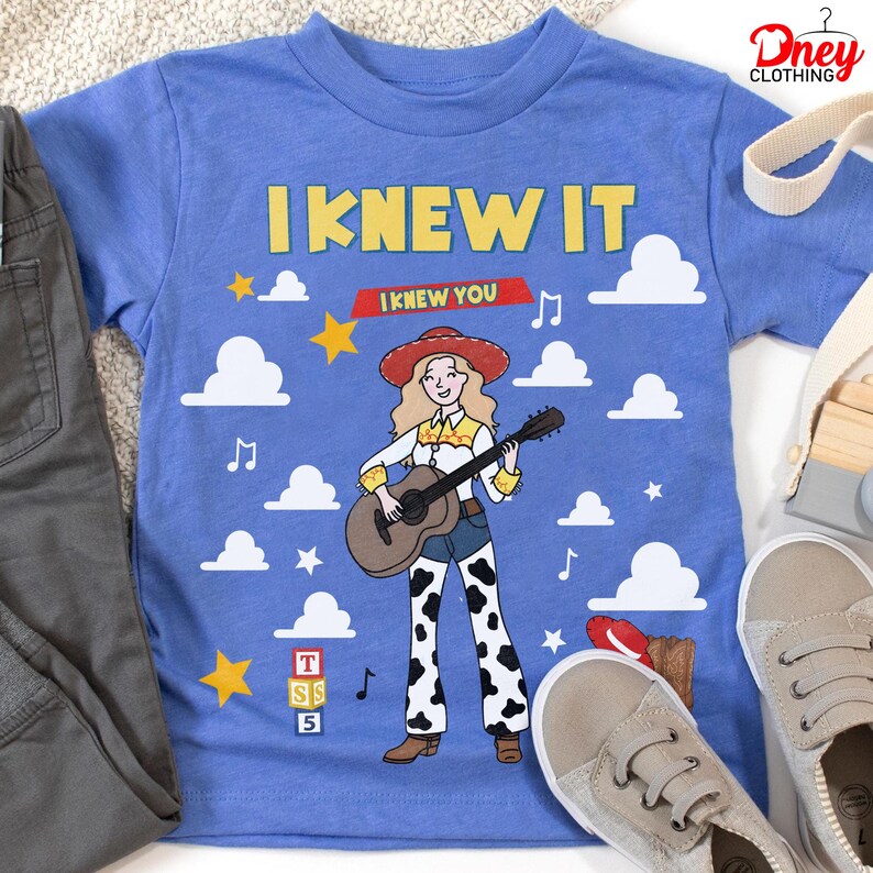May include: A blue t-shirt featuring a graphic of a cowgirl with long blonde hair, a red hat, and a white shirt with yellow accents, playing a brown acoustic guitar. The design includes white clouds, yellow stars, musical notes, and the text I KNEW IT.