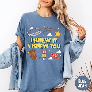Puede incluir: Una camiseta azul de manga corta con un estampado gr&aacute;fico que incluye el texto "I KNEW IT I KNEW YOU" y "infinity and beyond". El dise&ntilde;o presenta una pulsera de cuentas, una estrella amarilla, botas vaqueras marrones, bloques y un sombrero rojo.