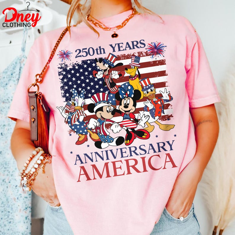 Puede incluir: Camiseta rosa claro con un dise&ntilde;o que celebra el 250 aniversario de Am&eacute;rica. El dise&ntilde;o presenta personajes de Disney y la bandera estadounidense. El texto dice "250th YEARS ANNIVERSARY AMERICA".