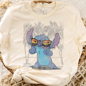 Op de afbeelding: Een cr&egrave;mekleurig t-shirt met korte mouwen en een afbeelding van een blauw alien-personage met een zwarte zonnebril waarin een zonsondergang en palmbomen weerspiegelen. De achtergrond toont een vervaagde grijze strandsc&egrave;ne met palmbomen.