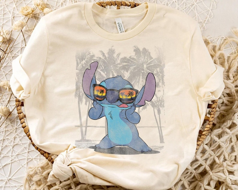 Op de afbeelding: Een cr&egrave;mekleurig t-shirt met korte mouwen en een afbeelding van een blauw alien-personage met een zwarte zonnebril waarin een zonsondergang en palmbomen weerspiegelen. De achtergrond toont een vervaagde grijze strandsc&egrave;ne met palmbomen.