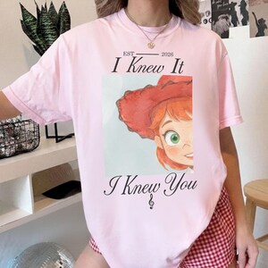 Puede incluir: Una camiseta de manga corta de color rosa claro con el gr&aacute;fico de un personaje de dibujos animados con pelo rojo y un sombrero de vaquero rojo. El texto dice EST 2026, I Knew It e I Knew You, con una nota musical bajo el texto.