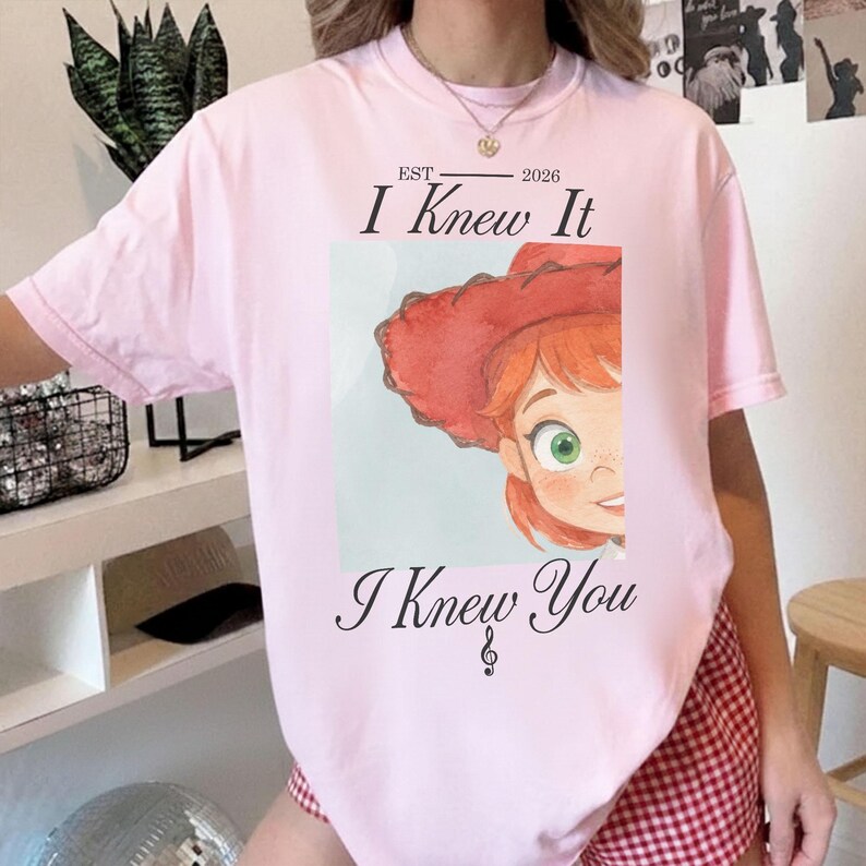 Puede incluir: Una camiseta de manga corta de color rosa claro con el gr&aacute;fico de un personaje de dibujos animados con pelo rojo y un sombrero de vaquero rojo. El texto dice EST 2026, I Knew It e I Knew You, con una nota musical bajo el texto.