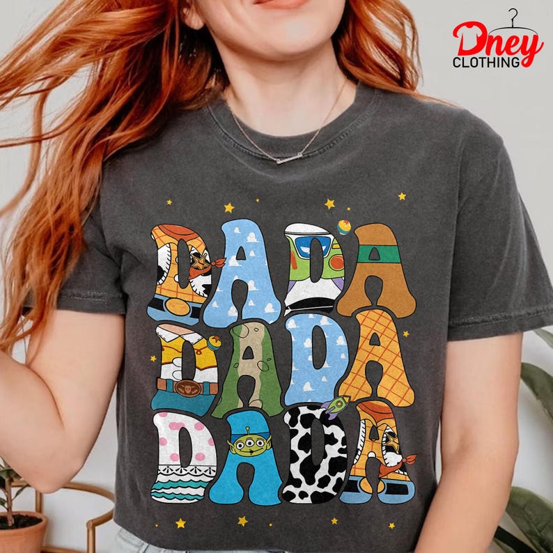 May include: Dark gray t-shirt with the word "DADA" repeated multiple times in colorful, patterned letters. The letters are designed with various cartoon character themes, including a cowboy boot and a space alien.