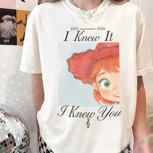 Puede incluir: Una camiseta de manga corta color crema con un estampado gr&aacute;fico de un personaje de dibujos animados con pelo rojo y un sombrero de vaquero rojo. El texto EST 2026 I Knew It I Knew You est&aacute; impreso en negro sobre la ilustraci&oacute;n.