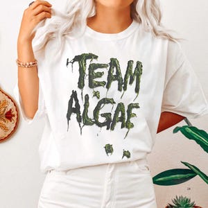 May include: A white short-sleeved t-shirt featuring the text "TEAM ALGAE" printed in a dark green, dripping, textured font that resembles slime or algae. The graphic is centered on the chest of the shirt, which has a crew neckline.