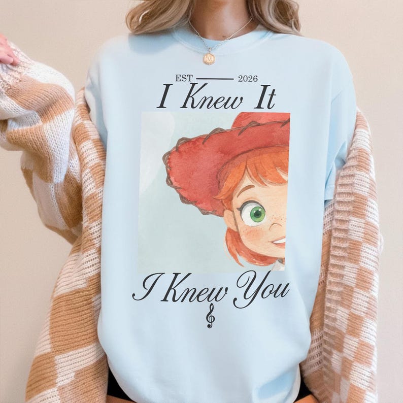 Puede incluir: Una sudadera de cuello redondo azul claro con un gr&aacute;fico de un personaje con sombrero rojo y ojos verdes. El texto dice EST 2026, I Knew It e I Knew You, con un s&iacute;mbolo de clave de sol debajo de la &uacute;ltima l&iacute;nea de texto en el frente.