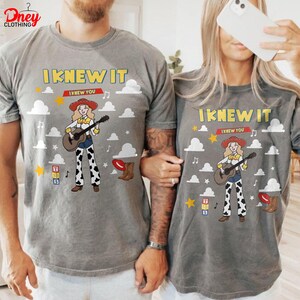 May include: A grey t-shirt featuring a cartoon cowgirl playing a guitar, with the text I KNEW IT I KNEW YOU, surrounded by white clouds, yellow stars, musical notes, alphabet blocks, and a brown cowboy boot, all printed in a graphic design.