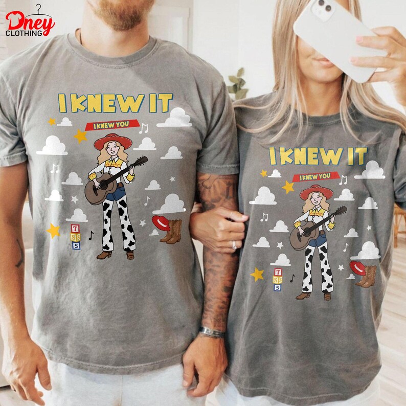 May include: A grey t-shirt featuring a cartoon cowgirl playing a guitar, with the text I KNEW IT I KNEW YOU, surrounded by white clouds, yellow stars, musical notes, alphabet blocks, and a brown cowboy boot, all printed in a graphic design.