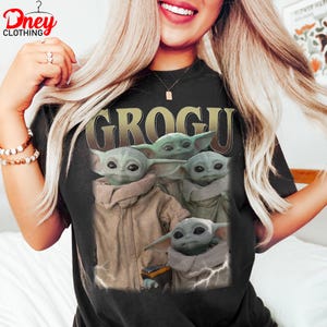 K&ouml;nnte beinhalten: Schwarzes T-Shirt mit einem Grogu-Motiv. Das Design zeigt mehrere Bilder von Grogu, auch bekannt als Baby Yoda, in verschiedenen Posen. Das Wort "GROGU" ist in goldfarbenen Buchstaben &uuml;ber den Bildern aufgedruckt. Das T-Shirt hat einen Rundhalsausschnitt.