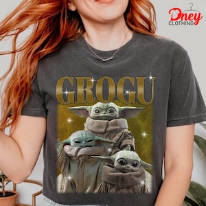 K&ouml;nnte beinhalten: Dunkelgraues T-Shirt mit dem Wort "GROGU" in goldfarbenen Buchstaben und einer Grafik von drei Grogu-Charakteren. Die Grogu-Charaktere sind hellgr&uuml;n mit gro&szlig;en Ohren. Das T-Shirt hat einen Vintage-Look.