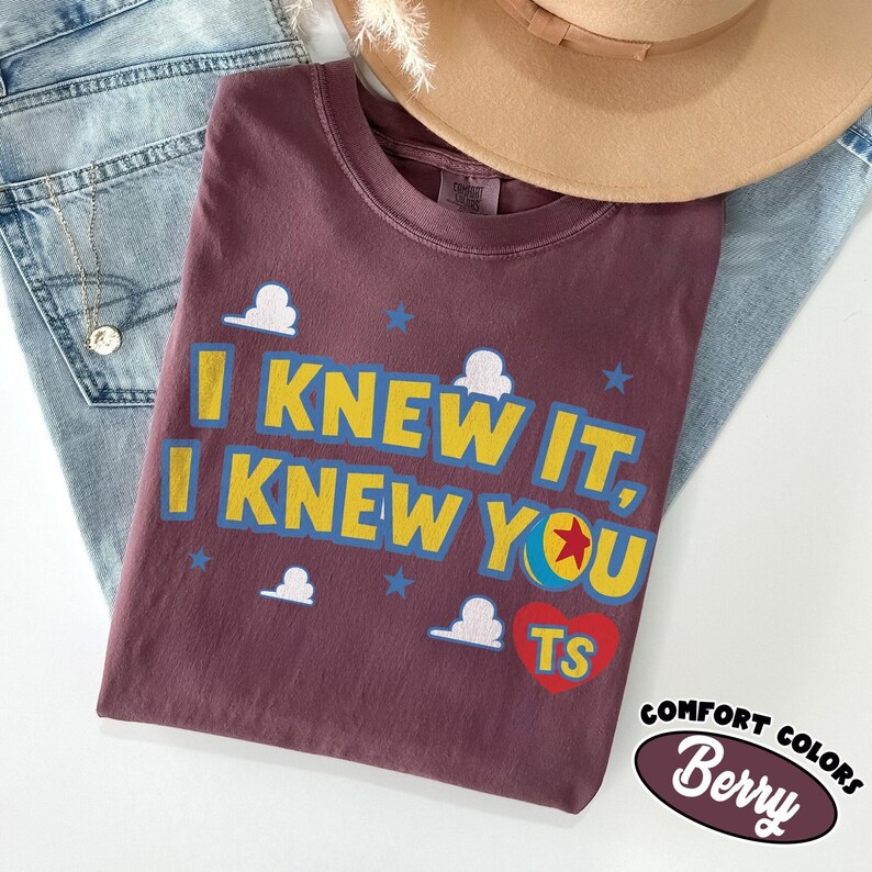 May include: A berry-colored t-shirt featuring the yellow text I KNEW IT, I KNEW YOU with a colorful ball graphic replacing the letter O. The design includes white cloud shapes, blue stars, and a red heart with the letters TS inside the heart.
