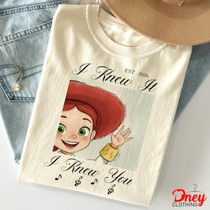 Op de afbeelding: Een cr&egrave;mekleurig t-shirt met een afbeelding van een stripfiguur met rood haar en een rode hoed die zwaait. De tekst "I Knew It EST-2026" staat boven de afbeelding en "I Knew You" met muzieknoten onder de afbeelding.