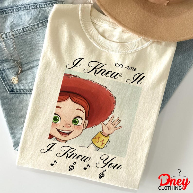 Op de afbeelding: Een cr&egrave;mekleurig t-shirt met een afbeelding van een stripfiguur met rood haar en een rode hoed die zwaait. De tekst "I Knew It EST-2026" staat boven de afbeelding en "I Knew You" met muzieknoten onder de afbeelding.