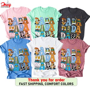 May include: A collection of six t-shirts in various colors, including pink, mint green, charcoal, blue, white, and mauve. Each shirt features the word "DADA" in a playful, colorful font with decorative patterns. The shirts are casual wear.