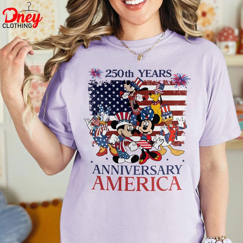 Puede incluir: Camiseta lila claro con un dise&ntilde;o de Mickey, Minnie, Donald y Pluto con atuendos patri&oacute;ticos. El dise&ntilde;o incluye una bandera americana y el texto "250th YEARS ANNIVERSARY AMERICA".