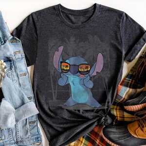 Op de afbeelding: Een donkergrijs T-shirt met korte mouwen en een afbeelding van het blauwe personage Stitch met een zonnebril waarin een strand bij zonsondergang met palmbomen wordt weerspiegeld. Het personage heeft grote oren en een speelse gezichtsuitdrukking.