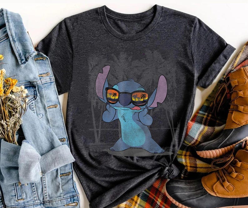 Op de afbeelding: Een donkergrijs T-shirt met korte mouwen en een afbeelding van het blauwe personage Stitch met een zonnebril waarin een strand bij zonsondergang met palmbomen wordt weerspiegeld. Het personage heeft grote oren en een speelse gezichtsuitdrukking.