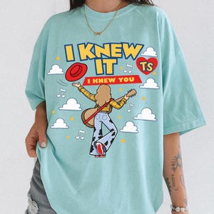 K&ouml;nnte beinhalten: Ein hellblaues T-Shirt mit einer Grafik einer Person mit langen Haaren, die eine Akustikgitarre spielt. Der Text lautet I KNEW IT und I KNEW YOU. Das Design umfasst einen roten Hut, ein rotes Herz mit TS, wei&szlig;e Wolken, Noten und gelbe Sterne.