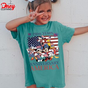 Puede incluir: Camiseta verde azulado con un dise&ntilde;o de Mickey Mouse, Minnie Mouse, Donald Duck y Pluto con atuendos patri&oacute;ticos. El dise&ntilde;o incluye una bandera estadounidense y el texto "250th YEARS ANNIVERSARY AMERICA". La camiseta es de Dney Clothing.