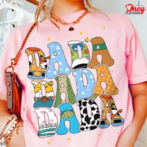 May include: Pink t-shirt with the word "DADA" in large, colorful letters. Each letter is designed with a different pattern and character theme, including a cowboy, a space ranger, and a cow print. The shirt has a crew neck and short sleeves.