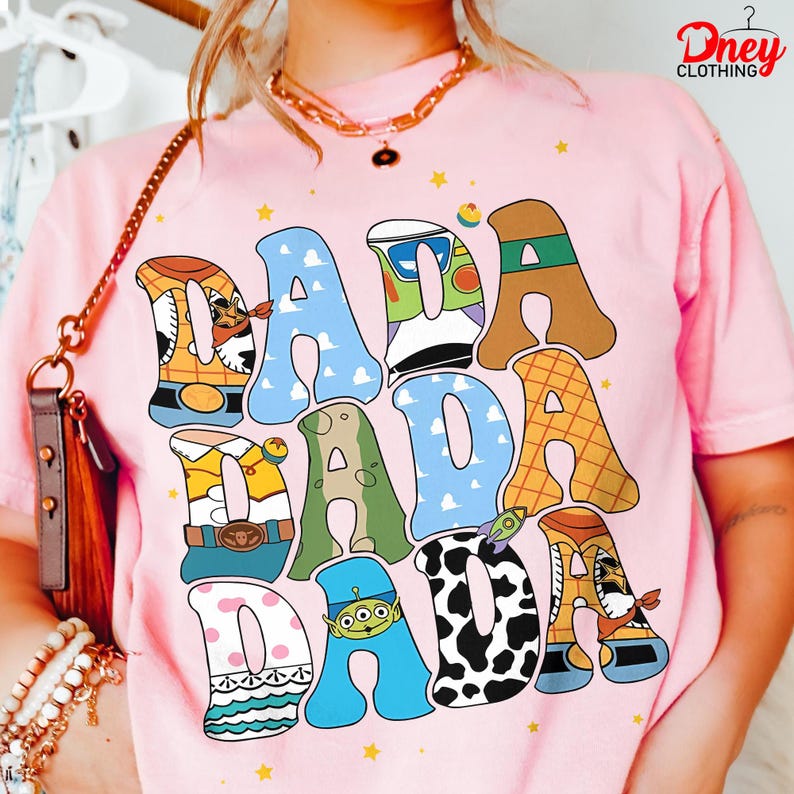 May include: Pink t-shirt with the word "DADA" in large, colorful letters. Each letter is designed with a different pattern and character theme, including a cowboy, a space ranger, and a cow print. The shirt has a crew neck and short sleeves.
