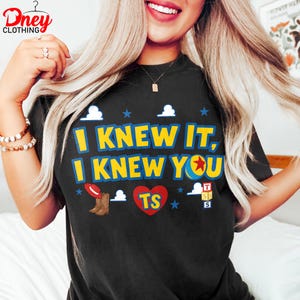 Puede incluir: Una camiseta negra con el texto amarillo "I KNEW IT, I KNEW YOU" y un coraz&oacute;n rojo que contiene las letras "TS". El dise&ntilde;o incluye gr&aacute;ficos de un sombrero de vaquero, botas marrones, una pelota con una estrella roja, bloques y nubes.