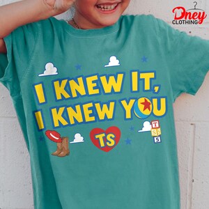 Puede incluir: Una camiseta color verde azulado con el texto amarillo I KNEW IT, I KNEW YOU. El dise&ntilde;o incluye un sombrero de vaquero rojo, un par de botas vaqueras marrones, un coraz&oacute;n rojo con las letras TS, una pelota azul y amarilla con una estrella roja y tres cubos.