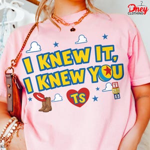 Puede incluir: Una camiseta rosa claro con el texto amarillo I KNEW IT, I KNEW YOU con bordes azules. El dise&ntilde;o incluye un coraz&oacute;n rojo con TS, un sombrero de vaquero rojo, botas marrones, una pelota azul y amarilla con una estrella roja, nubes y cubos.