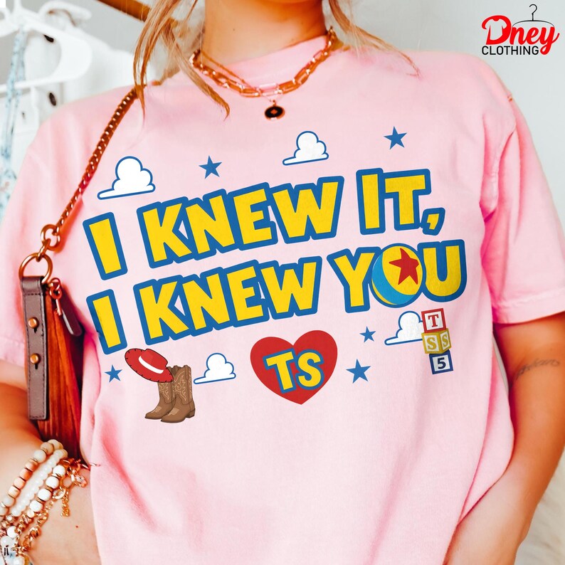 Puede incluir: Una camiseta rosa claro con el texto amarillo I KNEW IT, I KNEW YOU con bordes azules. El dise&ntilde;o incluye un coraz&oacute;n rojo con TS, un sombrero de vaquero rojo, botas marrones, una pelota azul y amarilla con una estrella roja, nubes y cubos.