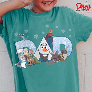May include: Teal t-shirt featuring the word "DAD" in a snowy blue design, with characters from the movie Frozen. The shirt includes images of Olaf, Elsa, Anna, Kristoff, Sven, and a small dragon, with snowflakes scattered around the design.