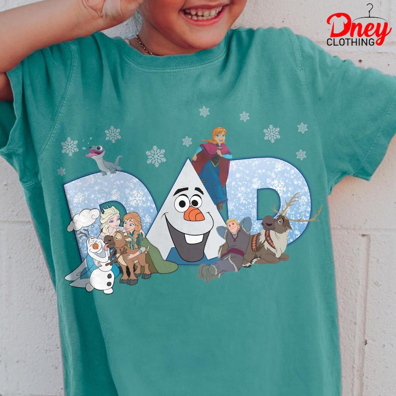 May include: Teal t-shirt featuring the word "DAD" in a snowy blue design, with characters from the movie Frozen. The shirt includes images of Olaf, Elsa, Anna, Kristoff, Sven, and a small dragon, with snowflakes scattered around the design.