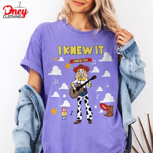 May include: A purple t-shirt featuring a graphic of a cowgirl with blonde hair, a red hat, and a guitar. The text reads I KNEW IT and I KNEW YOU. The design includes white clouds, yellow stars, musical notes, a red hat, brown boots, and three stacked blocks labelled T, S, and 5.