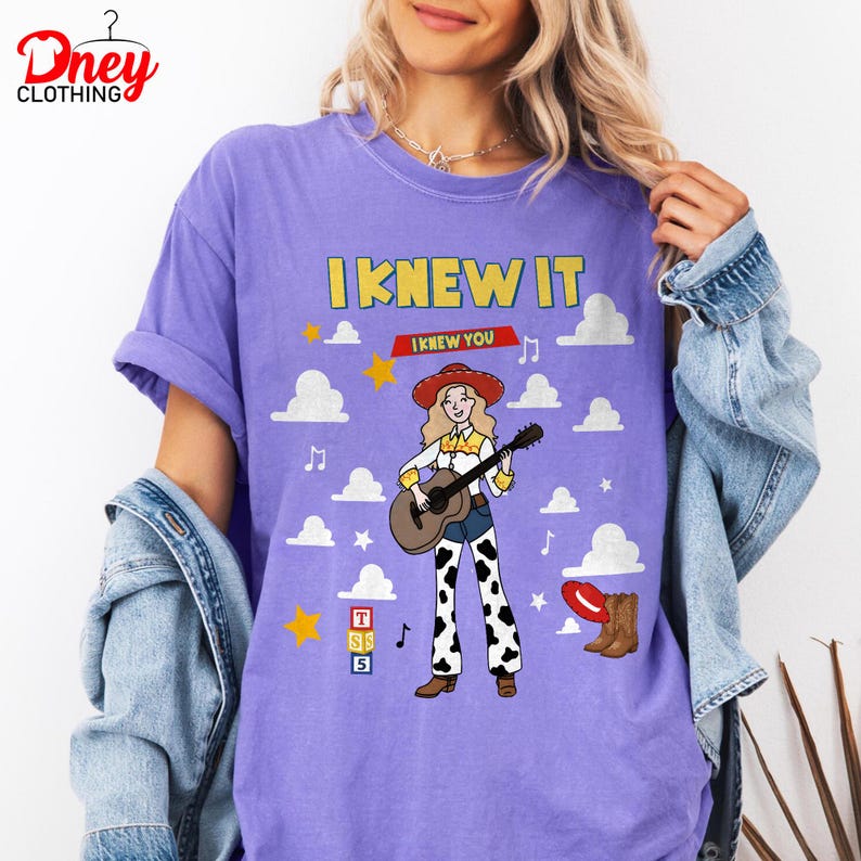 May include: A purple t-shirt featuring a graphic of a cowgirl with blonde hair, a red hat, and a guitar. The text reads I KNEW IT and I KNEW YOU. The design includes white clouds, yellow stars, musical notes, a red hat, brown boots, and three stacked blocks labelled T, S, and 5.