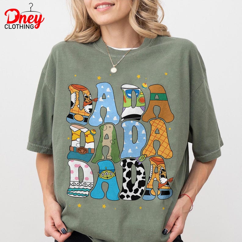 May include: Green t-shirt with the word "DADA" in large, colorful, cartoon-style letters. The letters are decorated with various toy-themed designs. The shirt has a relaxed fit and short sleeves.