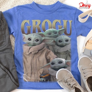 K&ouml;nnte beinhalten: Ein blaues T-Shirt mit einer Grafik von Grogu aus The Mandalorian. Das Wort "GROGU" ist in goldenen Buchstaben &uuml;ber dem Bild der Figur aufgedruckt. Das Shirt wird mit grauer Hose und beigen Turnschuhen kombiniert.