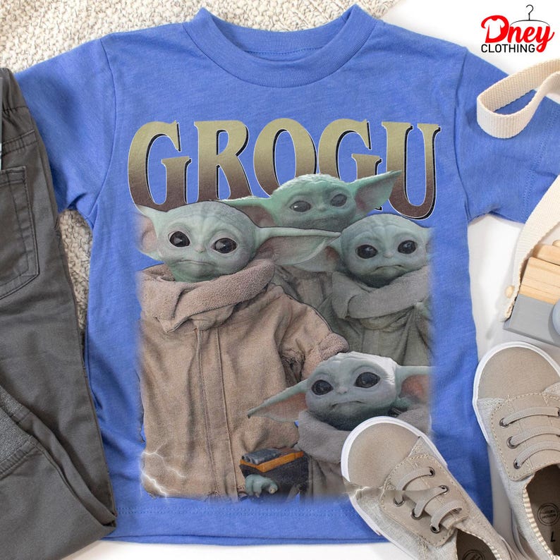 K&ouml;nnte beinhalten: Ein blaues T-Shirt mit einer Grafik von Grogu aus The Mandalorian. Das Wort "GROGU" ist in goldenen Buchstaben &uuml;ber dem Bild der Figur aufgedruckt. Das Shirt wird mit grauer Hose und beigen Turnschuhen kombiniert.