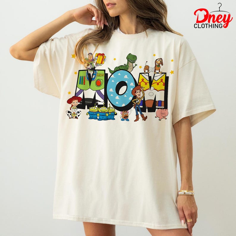May include: Cream-colored oversized t-shirt featuring the word "MOM" in colorful block letters with Toy Story characters. The "O" is blue with cloud details, and stars are scattered around the letters. The shirt has short sleeves.
