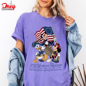 May include: A lilac t-shirt with a graphic of Mickey Mouse, Donald Duck, and Minnie Mouse in patriotic outfits, carrying a flag and drum. The shirt displays the text "250 Years of Freedom" and "United States Semiquincentennial".