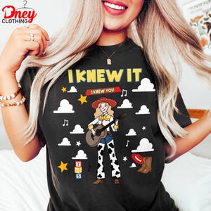 May include: A black t-shirt featuring a graphic of a blonde cowgirl character playing an acoustic guitar. The design includes the text "I KNEW IT" and "I KNEW YOU," white clouds, yellow stars, musical notes, a red hat, brown boots, and a stack of blocks.