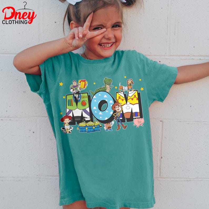 May include: Teal short-sleeved t-shirt with the word "MOM" in a colourful design featuring characters from the Toy Story film. The shirt has a crew neck and is made of a soft material. The Dney Clothing logo is in the top left corner.