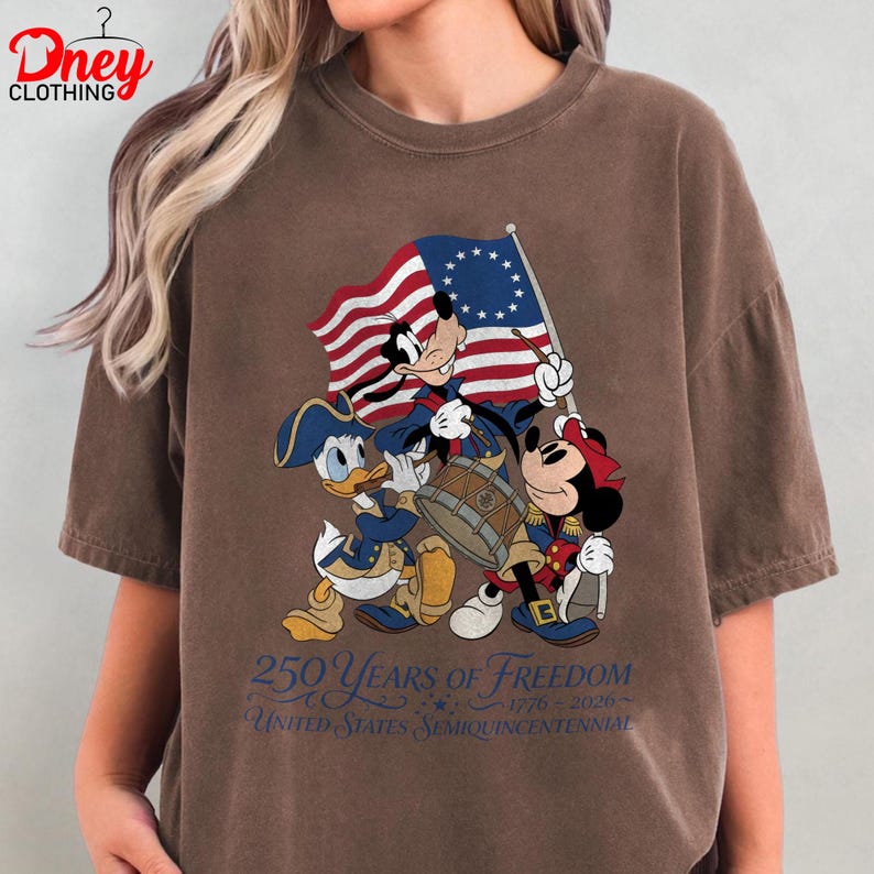 May include: Brown t-shirt with a patriotic design featuring Mickey Mouse, Donald Duck, and Minnie Mouse. The characters are shown with a flag and drum. Text reads "250 Years of Freedom" and "United States Semiquincentennial."
