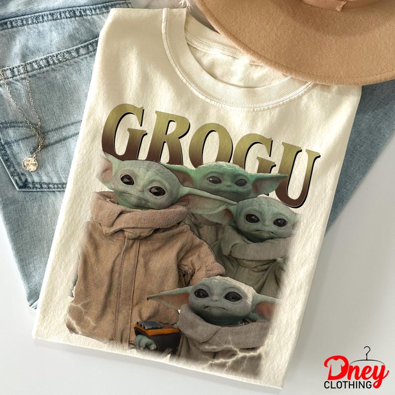 K&ouml;nnte beinhalten: Cremefarbenes T-Shirt mit dem Wort "GROGU" in einem braunen Farbverlauf. Das Shirt zeigt eine Grafik von vier Grogu-Charakteren. Das Shirt ist auf einer blauen Jeans gefaltet, mit einer Halskette und einem beigen Hut.