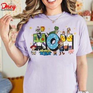 May include: A lilac t-shirt featuring the word "MOM" in a colourful cartoon design. The "M" is designed like Buzz Lightyear's suit, the "O" is blue with white clouds, and the other "M" is yellow. Toy Story characters surround the letters.
