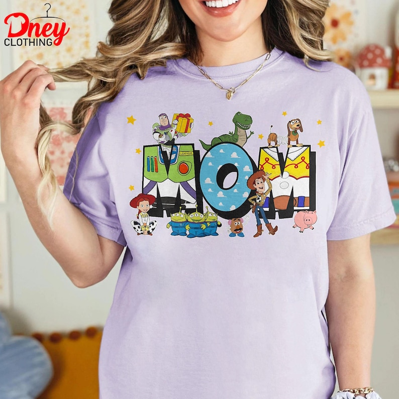 May include: A lilac t-shirt featuring the word "MOM" in a colourful cartoon design. The "M" is designed like Buzz Lightyear's suit, the "O" is blue with white clouds, and the other "M" is yellow. Toy Story characters surround the letters.