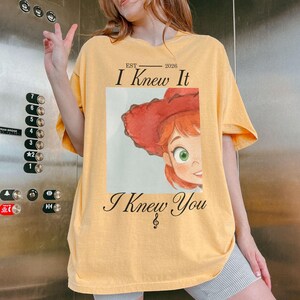 Puede incluir: Una camiseta de color amarillo claro con un estampado gr&aacute;fico de un personaje de dibujos animados con pelo rojo y un sombrero rojo. El texto EST 2026 I Knew It I Knew You est&aacute; impreso en negro sobre y bajo la imagen, con una nota musical.