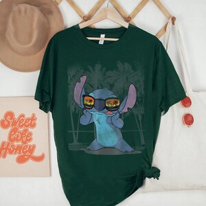 Op de afbeelding: Een donkergroen T-shirt met korte mouwen en een afbeelding van een blauw alien-personage met een zonnebril waarin een zonsondergang en palmbomen weerspiegelen, tegen een achtergrond van vage palmbomsilhouetten.